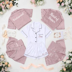 May include: A collection of bridal party pajamas in shades of pink and white. The pajamas include short-sleeved button-down shirts and shorts or pants. Some shirts are labeled "Bridesmaid" and "Maid of Honor". The name "Jane" is embroidered on some pieces.