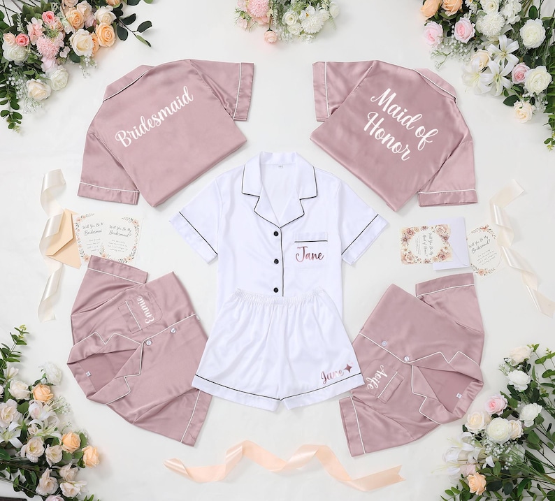 May include: A collection of bridal party pajamas in shades of pink and white. The pajamas include short-sleeved button-down shirts and shorts or pants. Some shirts are labeled "Bridesmaid" and "Maid of Honor". The name "Jane" is embroidered on some pieces.
