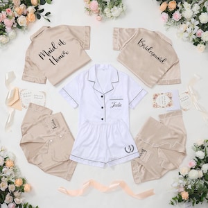 May include: A collection of bridal party pajamas in beige and white. The pajamas include short-sleeved button-down shirts and shorts. Some shirts are labeled "Maid of Honor", "Bridesmaid", and with names. The white set is personalized with the name "Jodie".