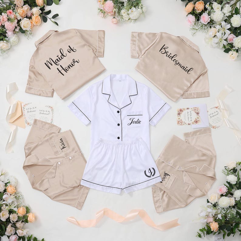 May include: A collection of bridal party pajamas in beige and white. The pajamas include short-sleeved button-down shirts and shorts. Some shirts are labeled "Maid of Honor", "Bridesmaid", and with names. The white set is personalized with the name "Jodie".