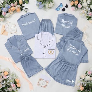 May include: A selection of light blue satin pyjama sets, comprising tops and shorts, with white piping. Some sets are labelled "Bridesmaid," "Maid of Honour," "Mother of the Bride," and "Emily" with a gold embroidered initial. The image is decorated with flowers and stationery.