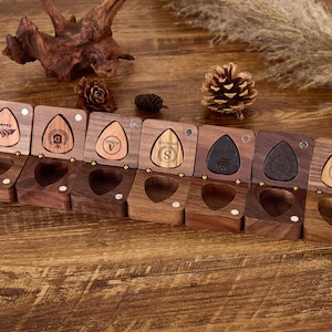 Puede incluir: Ocho cajas de anillos de madera con iniciales grabadas. Cada caja tiene una tapa con bisagras y un interior de terciopelo. Las cajas están dispuestas en una fila sobre una superficie de madera.