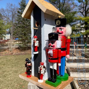May include: A wooden birdhouse with a light brown roof and a black and white striped front. The birdhouse is decorated with several toy nutcrackers in red, black, and white uniforms. The nutcrackers are of various sizes and are positioned on the birdhouse and its base.