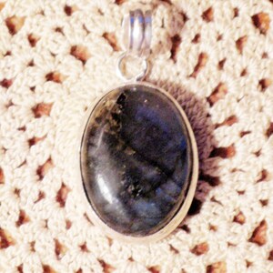 May include: A silver pendant with a dark blue oval labradorite gemstone. The pendant is on a white crocheted surface.