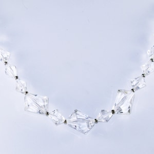May include: A clear crystal necklace displayed on a white bust. The necklace features a series of faceted, geometric crystal beads strung together with small silver-colored connectors. The crystals vary in size and shape, creating a sparkling effect.
