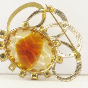 May include: A gold tone brooch featuring a large oval amber stone set within a circle of clear rhinestones. The brooch has a decorative design of interlocking circles.
