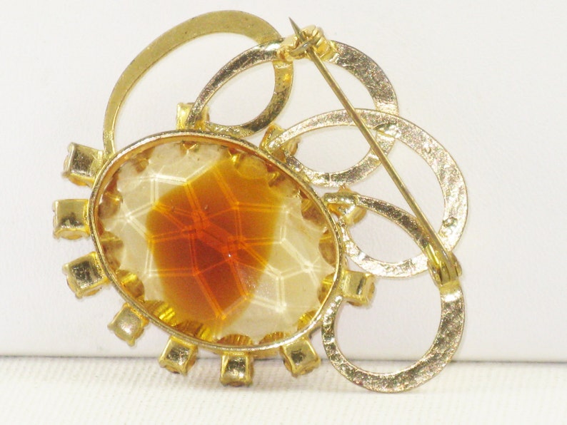 May include: A gold tone brooch featuring a large oval amber stone set within a circle of clear rhinestones. The brooch has a decorative design of interlocking circles.