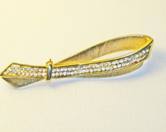 May include: A gold-toned brooch in the shape of a bow with clear rhinestones. The bow is made of two pieces of metal that are joined together in the middle. The rhinestones are set in a row along the edge of each piece of metal.