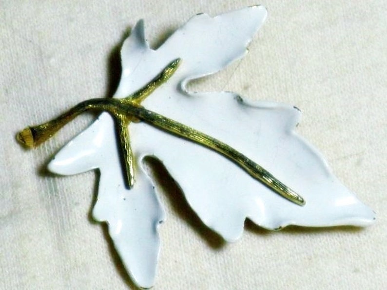 May include: White enamel leaf brooch with a gold stem and veins. The leaf is a simple shape with a slightly pointed tip.
