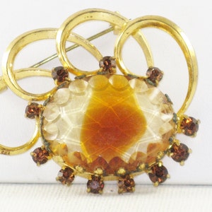 May include: A gold tone brooch featuring a large, oval, carved, amber-coloured glass stone surrounded by smaller, brown glass stones. The brooch is made of four interlocking circles.