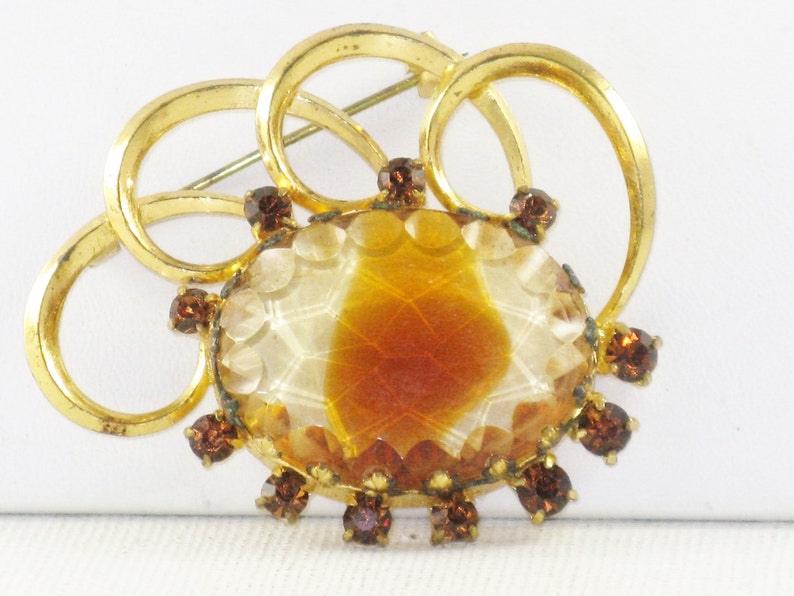May include: A gold tone brooch featuring a large, oval, carved, amber-coloured glass stone surrounded by smaller, brown glass stones. The brooch is made of four interlocking circles.