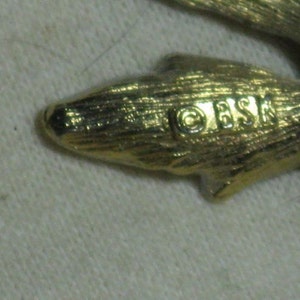 May include: Close-up of a gold-toned metal leaf-shaped brooch with the inscription "OESK" on the back.