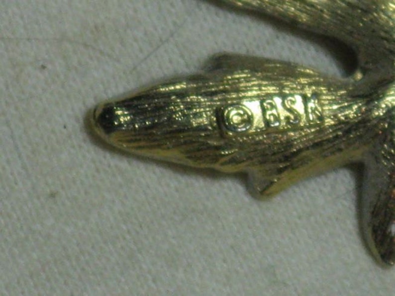 May include: Close-up of a gold-toned metal leaf-shaped brooch with the inscription "OESK" on the back.