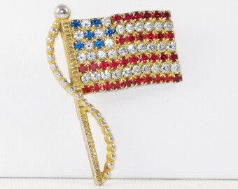 May include: A gold-tone brooch in the shape of an American flag. The flag is made of red, white, and blue rhinestones.