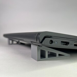 May include: A gray laptop stand made of plastic with a triangular shape. The stand is designed to elevate a laptop for better ergonomics and airflow.