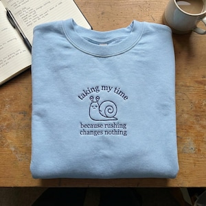 May include: A light blue sweatshirt with the embroidered phrase "taking my time" above a snail illustration, and "because rushing changes nothing" below. The sweatshirt is folded on a wooden surface, with a notebook and a mug in the background.