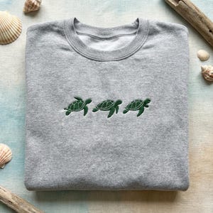 Könnte beinhalten: Ein hellgraues Sweatshirt mit drei gestickten grünen Meeresschildkröten auf der Brust. Das Sweatshirt mit Rundhalsausschnitt ist sauber gefaltet, mit Muscheln und Treibholz im Hintergrund.