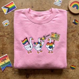 May include: A light pink crewneck sweatshirt featuring an embroidered design of three white geese. One goose holds a rainbow flag, another wears a rainbow scarf and holds a megaphone with the text HONK!, and the third holds a rainbow heart and a sign reading PRIDE IS PROTEST.