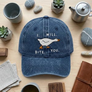 May include: A denim blue baseball cap with the embroidered words "I. WILL. BITE. YOU." and a white goose graphic with orange feet and beak. The hat is surrounded by decorative items.