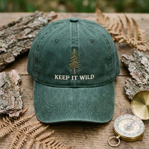 May include: A green baseball cap with the embroidered phrase "KEEP IT WILD" and a tree graphic. The cap is made of a soft, textured material and is displayed on a wooden surface with natural elements.