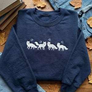 May include: A navy blue sweatshirt with white embroidered foxes and butterflies. The foxes are arranged in a row across the chest. The sweatshirt is made of a soft material and is suitable for casual wear.