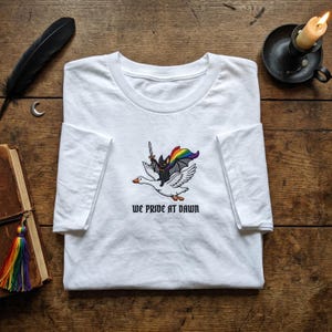 May include: A white t-shirt featuring an embroidered graphic of a black bat wearing a rainbow cape and holding a sword while riding a white goose. Below the graphic, the text reads WE PRIDE AT DAWN in a black, stylized, gothic-inspired font.