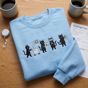 Könnte beinhalten: Hellblaues Sweatshirt mit gesticktem Design von schwarzen Katzen zum Thema Radiologie. Die Katzen sind mit Röntgengeräten, Schutzbrillen und Strahlenausweisen dargestellt. Das Sweatshirt wird mit Kaffeetassen und einem Radiologie-Vorbereitungsbogen präsentiert.