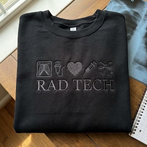 May include: Black crew neck sweatshirt with embroidered medical symbols and the text "RAD TECH" in grey. The design includes a rib cage, ultrasound, heartbeat, syringe, and bow. The sweatshirt is on a wooden surface.