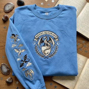 May include: A pale blue sweatshirt with dragon embroidery on the sleeve and chest. The chest design includes a dragon and the words "RIDERS QUADRANT". The sleeve shows multiple dragons with clouds. The sweatshirt is on a wooden surface.