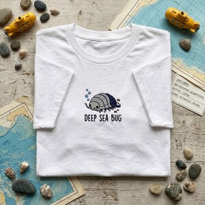 May include: A white short-sleeved t-shirt featuring an embroidered graphic of a grey and dark blue deep sea isopod with small blue bubbles and the text DEEP SEA BUG printed in black capital letters centred on the chest area of the garment.