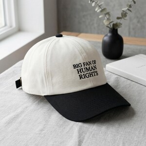 May include: A cream and black baseball cap featuring the text "BIG FAN OF HUMAN RIGHTS". The cap has a curved peak and an adjustable strap at the back.