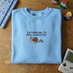 May include: Light blue sweatshirt with the text "I'M STARTING TO FEEL EXHAUSTED." and a pixelated snail and mushroom design. The sweatshirt is a crew neck style and is made of a soft material. The design is in shades of brown, green, and red.