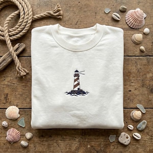 Könnte beinhalten: Ein cremefarbenes Sweatshirt mit Rundhalsausschnitt und einer kleinen, mittigen Stickerei eines Leuchtturms mit braun-weißen Diagonalstreifen, einem dunkelblauen Dach und dunkelblauen Wellen am Fuß, platziert auf einer rustikalen Holzoberfläche mit Muscheln.