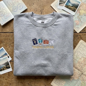 May include: A heather gray sweatshirt with embroidered travel-themed icons: a passport, globe, camera, and airplane. The words "TRAVEL LOVER" are embroidered below the icons in gold thread. The sweatshirt is a crew neck style.