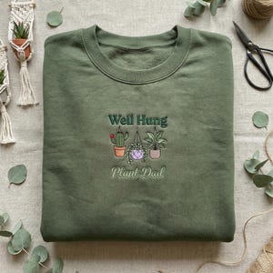 May include: Olive green sweatshirt with embroidered text and plant illustrations. The text reads "Well Hung Plant Dad" above three hanging plants and a cactus. The sweatshirt is folded neatly on a neutral surface.