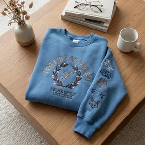 May include: A light blue sweatshirt with embroidered text and graphics. The front features "FOXFIRE ACADEMY" in a circular design with a letter "F" and the text "KEEPER OF THE LOST CITIES". The sleeve has embroidered animal designs.