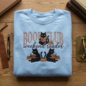 May include: A pale blue sweatshirt with the embroidered text "BOOK CLUB weekend reader". The design features three black cats reading books. The top cat is sitting on a stack of books, and the other two are sitting on either side of the text.
