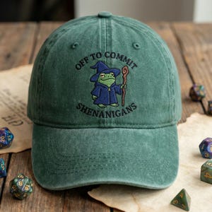 May include: Green baseball cap with the embroidered phrase "OFF TO COMMIT SHENANIGANS" and a cartoon frog wizard. The frog is wearing a blue robe and hat, holding a staff. The cap is on a wooden surface with dice.