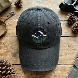 May include: A black baseball cap with a circular patch featuring a mountain scene with white, blue, and green embroidery. The cap is made of a worn-looking fabric and has a curved brim. The hat is a classic, casual accessory.