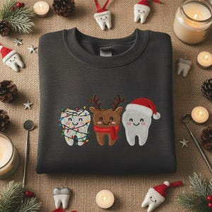 May include: Dark gray sweatshirt with embroidered Christmas teeth designs. One tooth is decorated with Christmas lights, another is a reindeer, and the third is wearing a Santa hat. The sweatshirt is surrounded by Christmas decorations.
