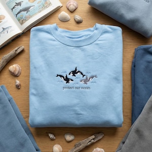 Könnte beinhalten: Hellblaues Sweatshirt mit gestickten Orcas und dem Text "protect our ocean". Das Bild zeigt Muscheln, ein Buch mit Meereslebewesen und weitere blaue Sweatshirts.
