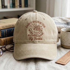 May include: Beige baseball cap featuring the embroidered text "Easily Distracted By Books" and a duck with books design. The cap has a curved peak and a button on top. Books and a mug are visible in the background.