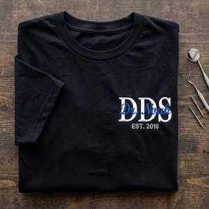 May include: Black t-shirt featuring the text "DDS Dr. Noah EST. 2016" in white and blue. Dental instruments are placed on a wooden surface.