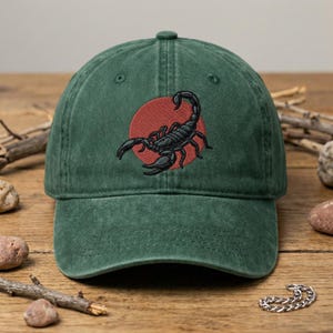 May include: A forest green baseball cap featuring an embroidered design on the front. The design consists of a solid red circle with a detailed black scorpion positioned in the center. The cap has a structured crown and a curved brim.