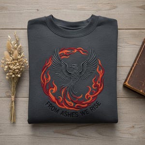 May include: Dark gray sweatshirt featuring an embroidered phoenix design in black, red, and orange flames. The text "FROM ASHES WE RISE" is embroidered below the phoenix. The sweatshirt is made of a soft material.
