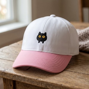 May include: A white baseball cap with a pink peak. The cap has an embroidered black cat with yellow eyes on the front. The hat is on a wooden surface.