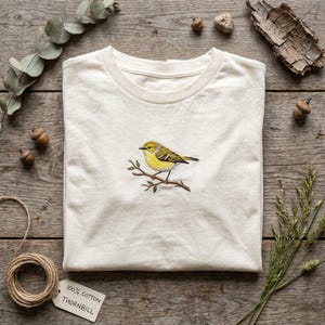 Pu&ograve; includere: T-shirt color crema con un ricamo di un uccello giallo appollaiato su un ramo. L'uccello ha dettagli verdi e gialli. La maglietta &egrave; in 100% cotone e ha la scritta "THORNBILL" sull'etichetta.