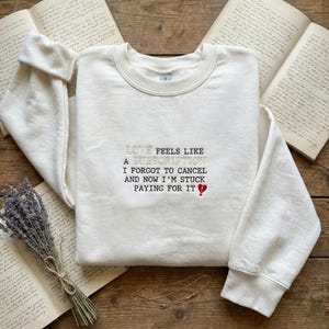 May include: Cream-colored sweatshirt with the embroidered text "LOVE FEELS LIKE A SUBSCRIPTION I FORGOT TO CANCEL AND NOW I'M STUCK PAYING FOR IT" in black and a red heart symbol. The sweatshirt is on a wooden surface with open books and a lavender bundle.