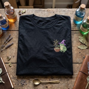 May include: Black t-shirt with an embroidered design of a green frog wearing a purple wizard hat, stirring a potion in a brown cauldron. The design includes gold stars and a wand. The shirt is folded on a wooden surface with potion bottles.