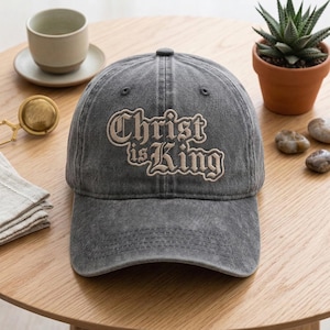 Könnte beinhalten: Graue Baseballkappe mit dem gestickten Text "Christ is King" in einer stilisierten Schriftart. Die Kappe hat einen gebogenen Schirm und einen Used-Look. Der Hut liegt auf einer Holzoberfläche.